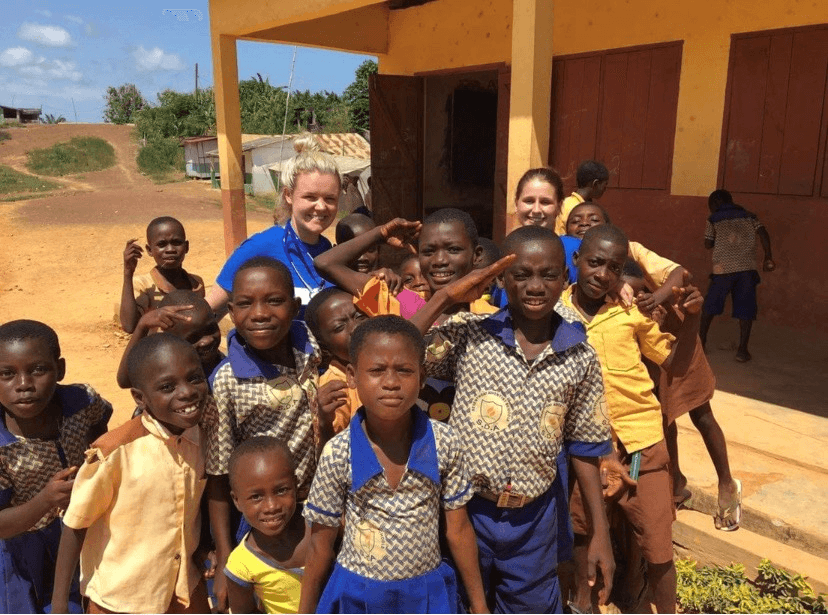 Group of Ghanaian children with Courtney and Emily together outside a building, with some wearing school uniforms.