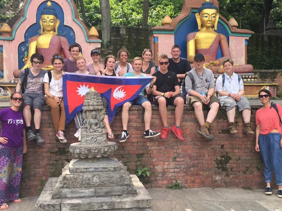 Group of students posing with a Nepalese flag in front of traditional Buddha statues.