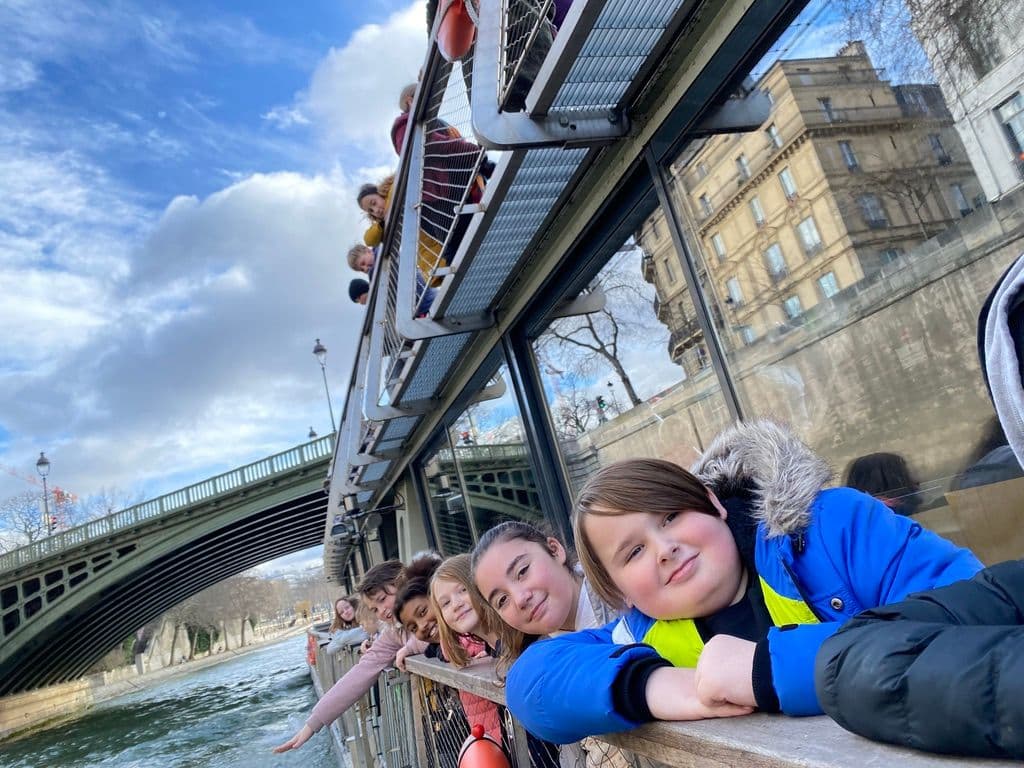 Children leaning on a boat railing on a river boat with a bridge in the background, enjoying a day out.