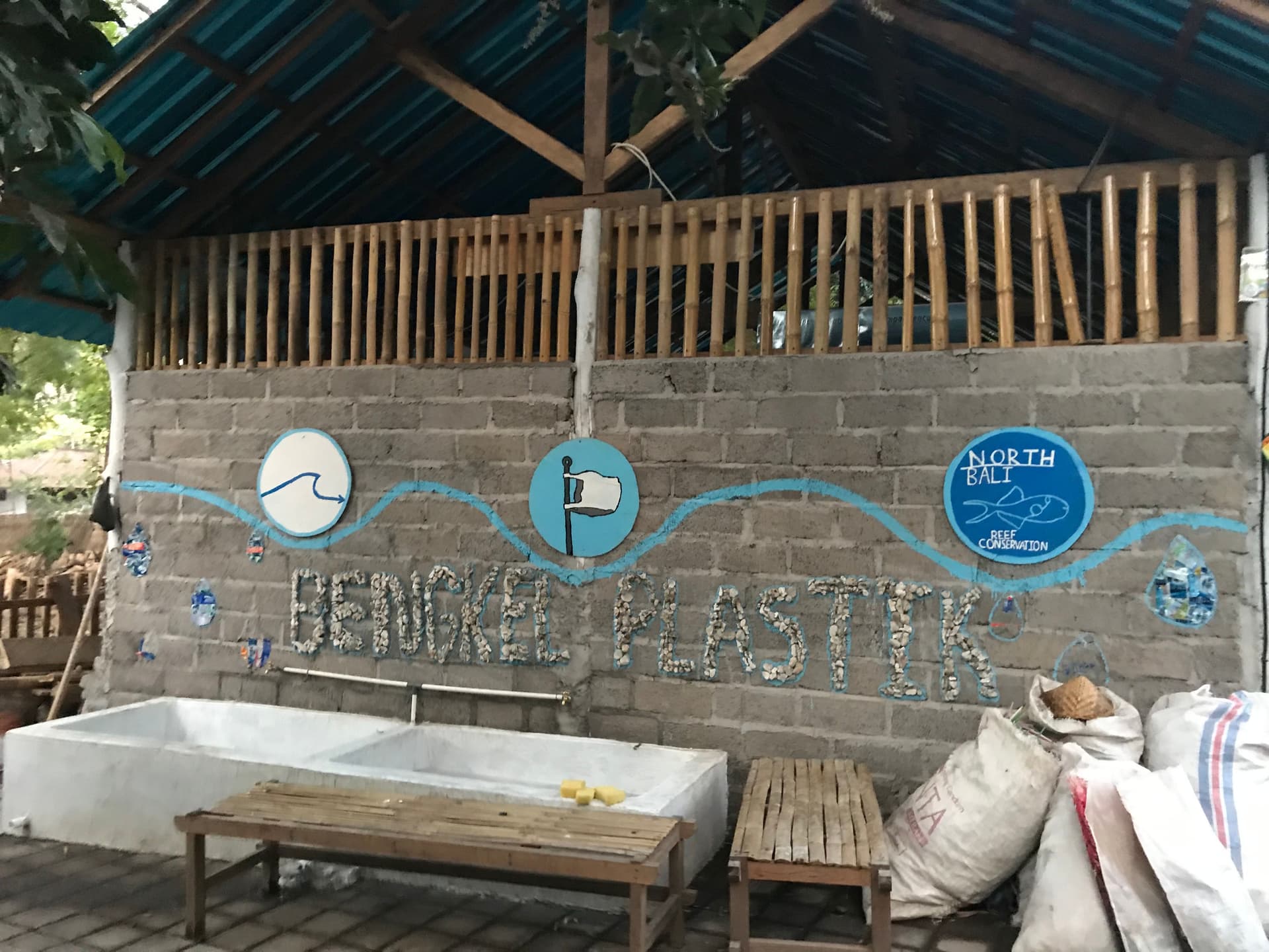 A wall with an environmental message, "end plastic", painted on it, with illustrations representing ocean life and plastic pollution, located in an outdoor setting with sinks and bags in the foreground.