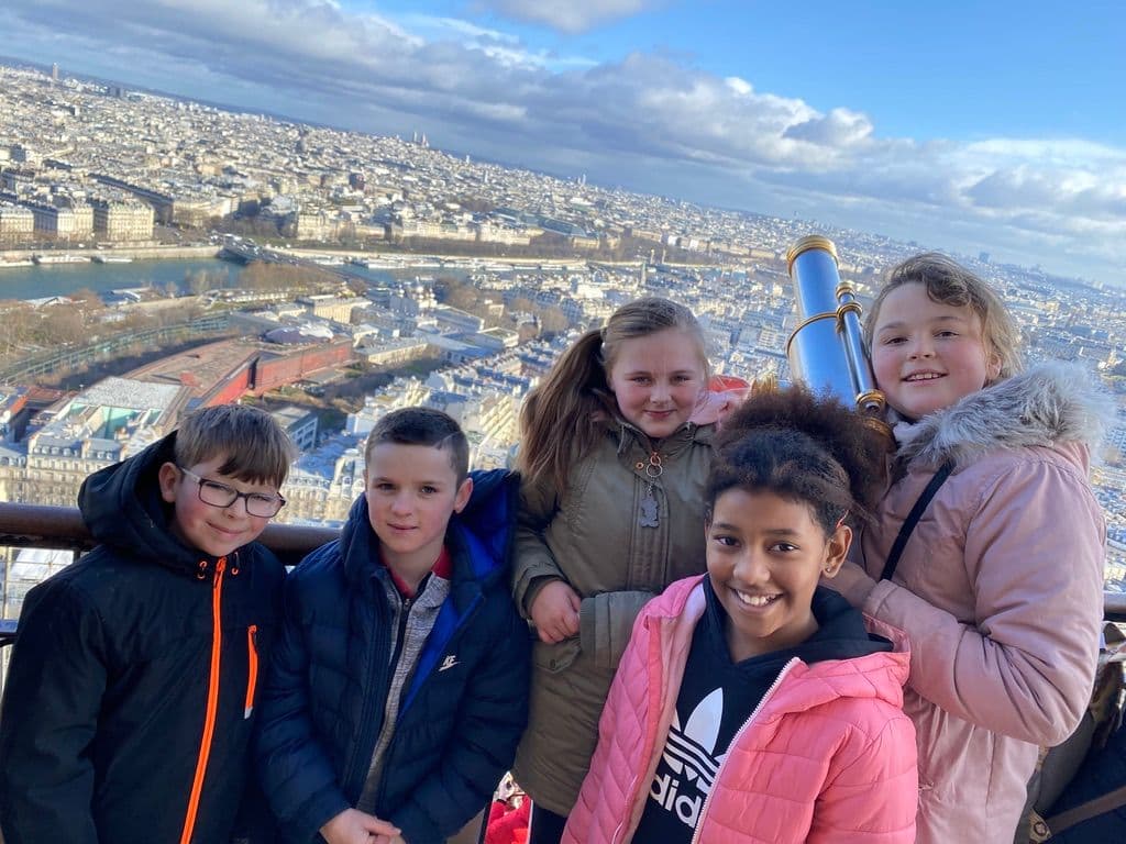 Group of children posing for a photo with a scenic cityscape view of Paris from an observation deck.